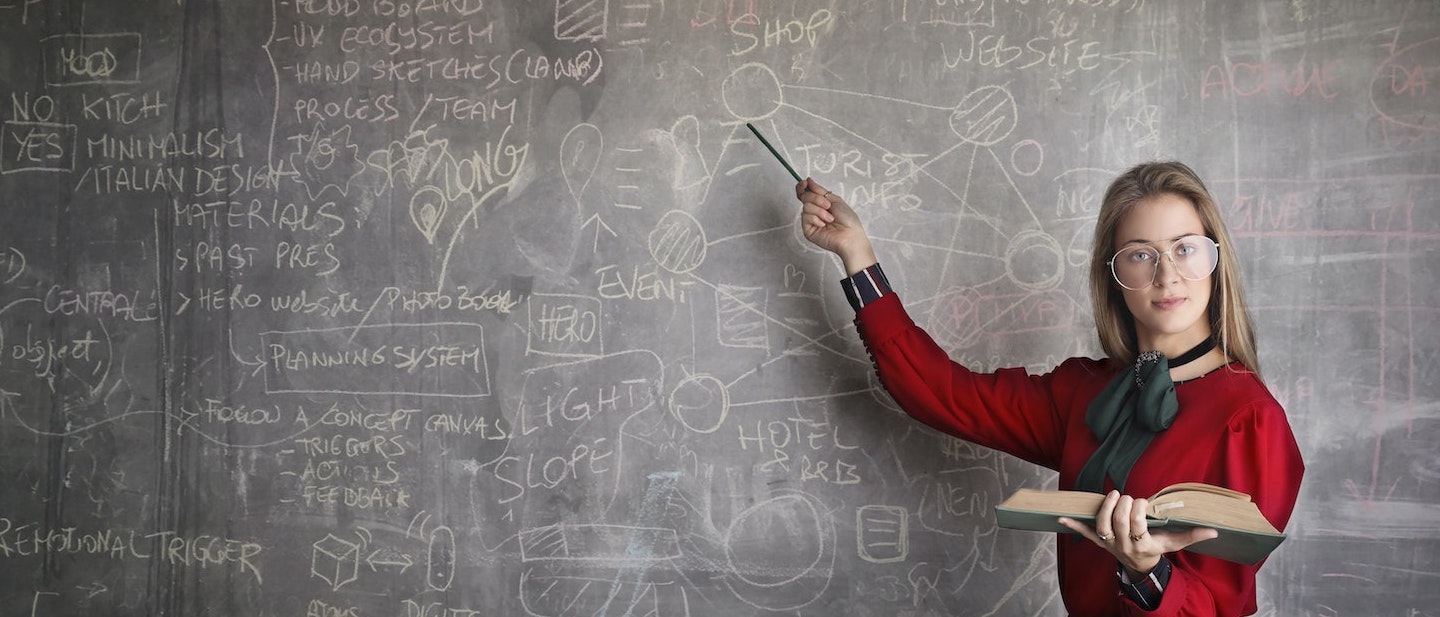 Foto: Lehrerin mit Buch zeigt auf Tafel